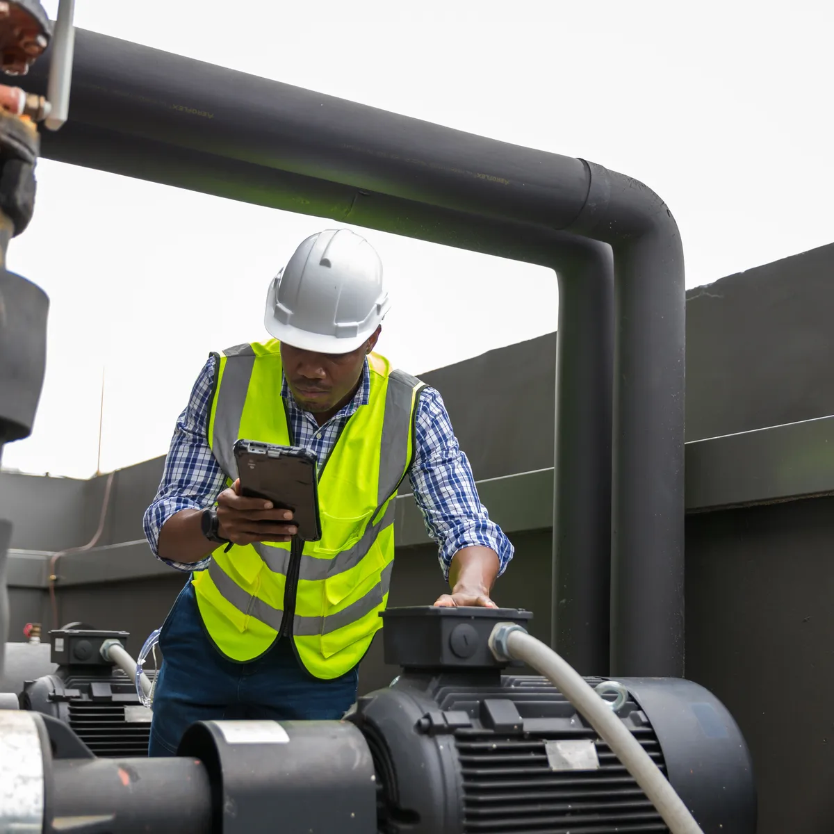 Worker in a hard hat and safety vest using a tablet beside pumps and piping on a commercial rooftop